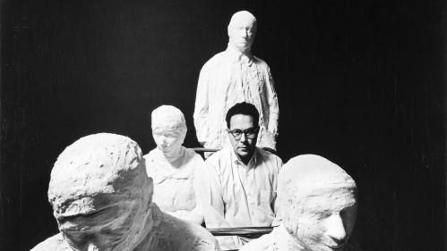 George Segal sits amongst plaster casts of bus riders. George is staring directly at the camera which is eye-level with him.